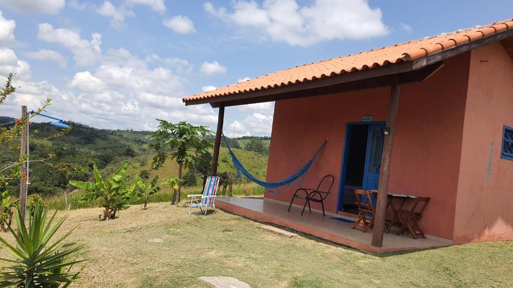 Casa rústica da pousada Sitio Aconchego Verde Guararema com paredes laranja, uma janela e porta, além de uma varanda com cadeiras e rede azul. O lugar é rodeado de natureza e tem vista para as montanhas