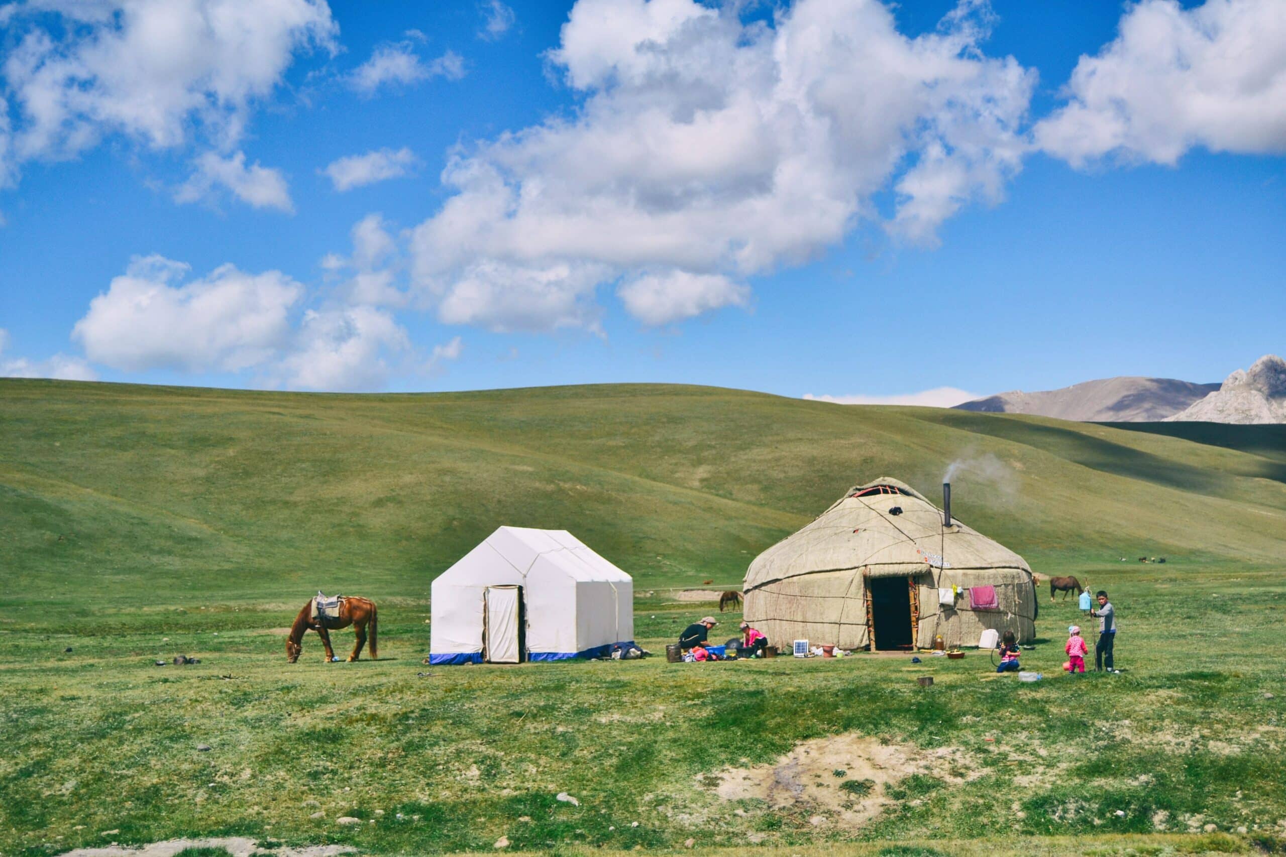 Vista de duas tendas em Song-Kul, Quirguistão, no meio das montanhas verdes durante o dia, com um cavalo no pasto e uma família a frente das tendas.
