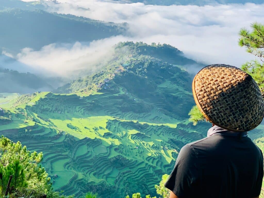 Pessoa usando uma blusa cinza e chapéu típico no formato de triangulo, de costas, olhando as montanhas verdes que são os Terraços de arroz de Banaue. Há nuvens entre as montanhas ao fundo