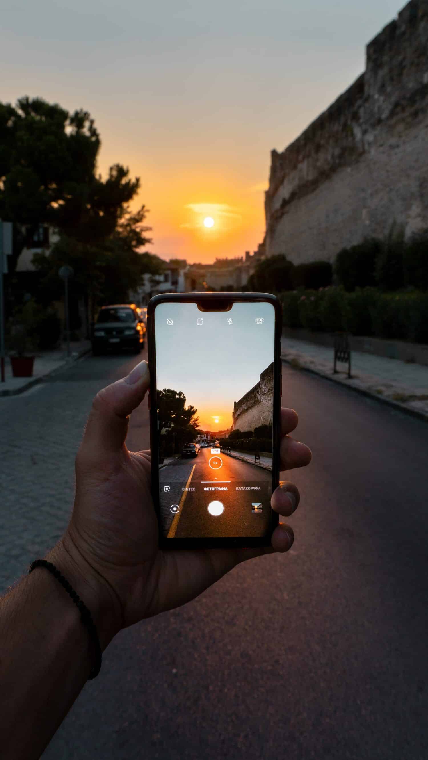 Uma mão segurando um celular tirando foto de uma rua de Tessalônica, na Grécia, com um pôr do sol alaranjado ao fundo. Imagem para ilustrar o post chip de celular Grécia