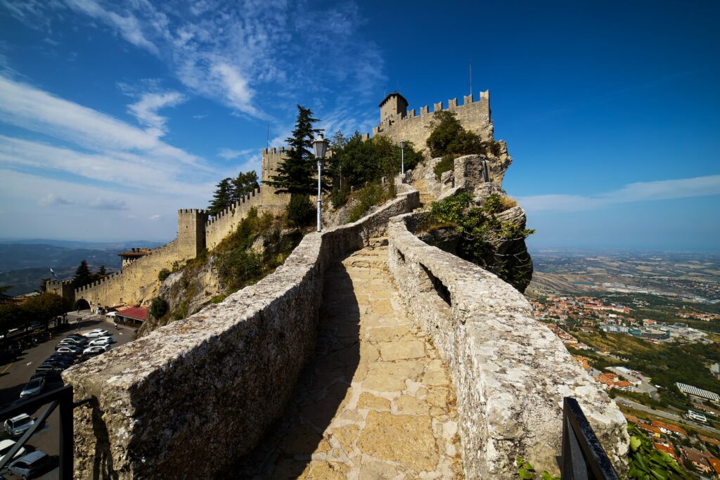 Caminho com chão e parede de pedras dando acesso a um castelo histórico de San Marino. Ao fundo, há uma vista panorâmica da cidade e, no lado esquerdo, um pouco mais abaixo, tem um estacionamento com vários carros parados