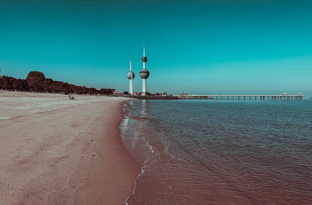 Vista da Torres Kuwait ao fundo durante o dia com mar e areia a frente do monumento.