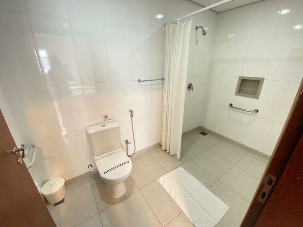Banheiro adaptado do Valinhos Plaza Hotel com barras de apoio perto da privada e dentro do box que é de cortinas, o espaço é amplo