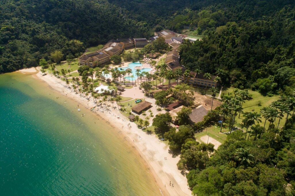 Vista aérea do Vila Galé Eco Resort Angra com muitas árvores e montanhas cercando a propriedade e, na frente do hotel, há uma praia de mar verde claro e uma faixa de areia clara, é possível ver também as piscinas do local, para representar resorts em Angra dos Reis