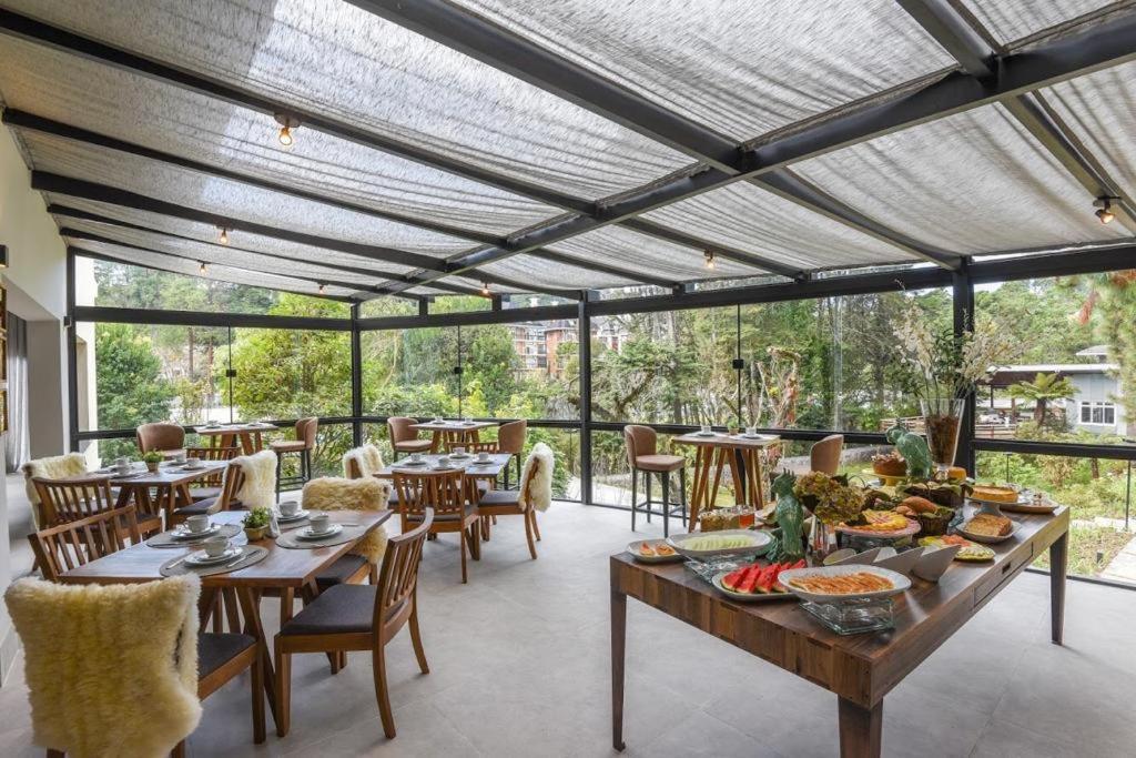 Área de refeições do Villa Amistà Campos do Jordão com tudo cercado por janelas de vidro dando vista para muitas árvores, do lado de dentro há mesas e cadeiras de madeira