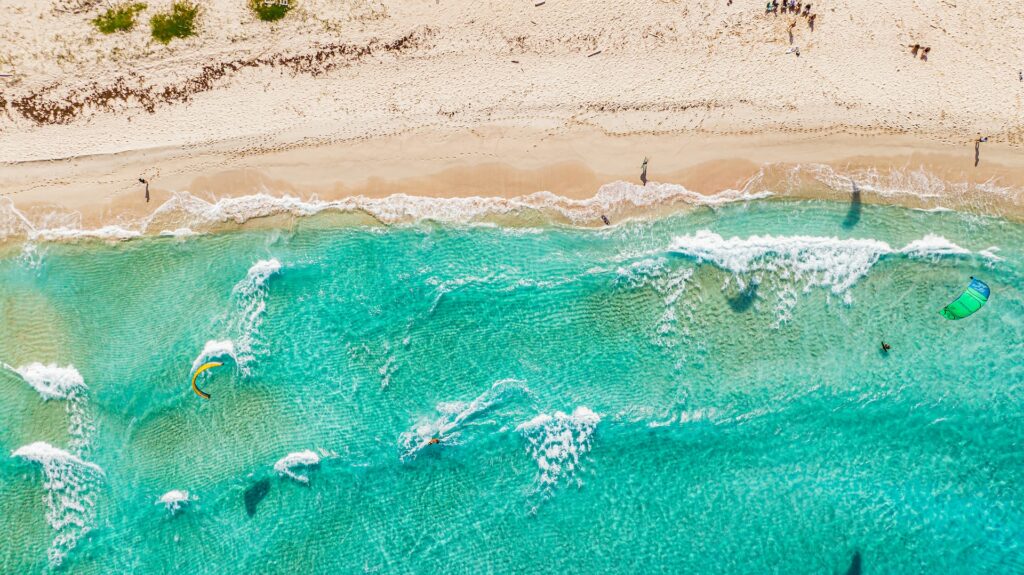 Vista aérea do mar verde-água, pessoas praticando kite surf, e uma faixa de areia clara na parte de cima da foto, ilustrando o post de chip celular Aruba
