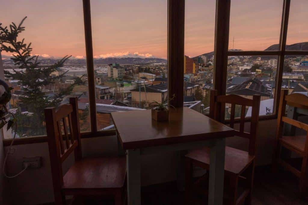 restaurante da Hostería Aonikenk com janelas amplas e vista para o por do sol. Na imagem há uma mesa com duas cadeiras e um céu rosada ao fundo