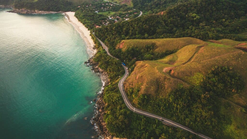 Vista de cima da praia de Toque Pequeno, São Sebastião, com mar azul, pequena faixa de areia, areia verde e estrada.