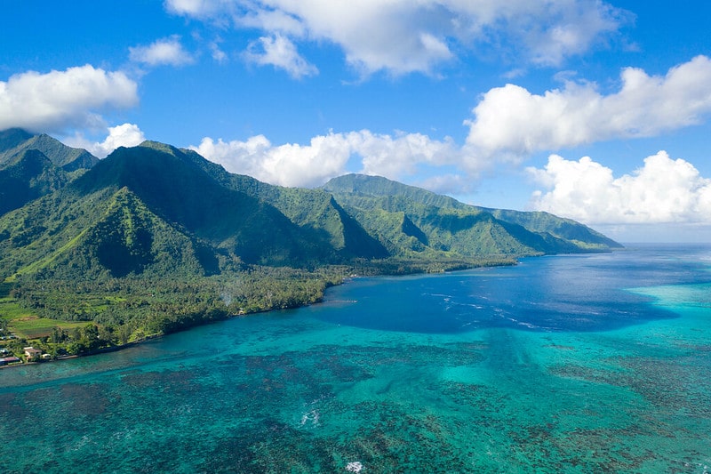 Vista do mar de Tahiti, com montanhas no lado direito e bastante natureza verde, além do céu ensolarado com algumas nuvens brancas