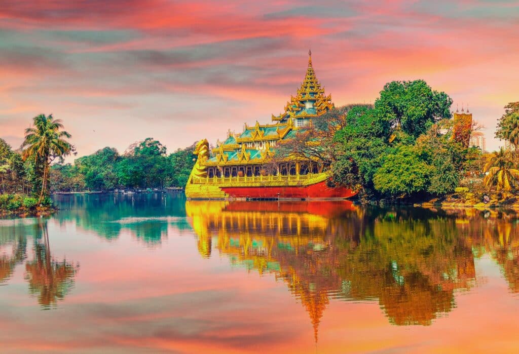 Construção asiática, similar a uma pirâmide em um barco, num lago, com o céu em tons laranja e rosa refletindo na água. Há bastante natureza nas redondezas