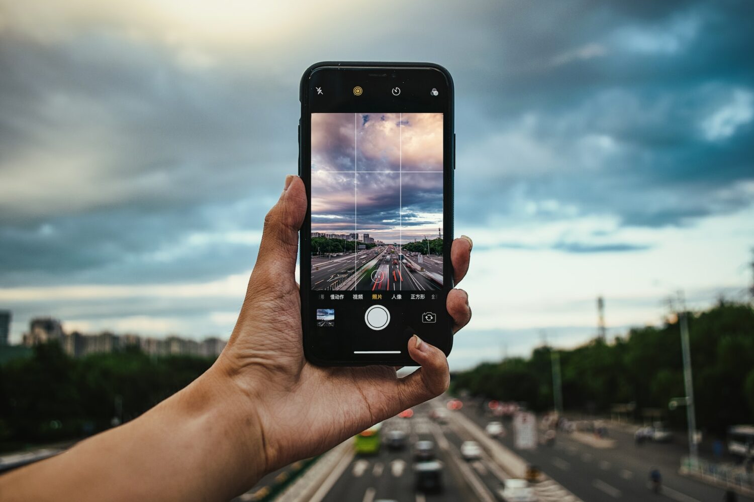 Uma mão branca segurando um celular na vertical para fotografar uma avenida, para representar chip celular Pequim