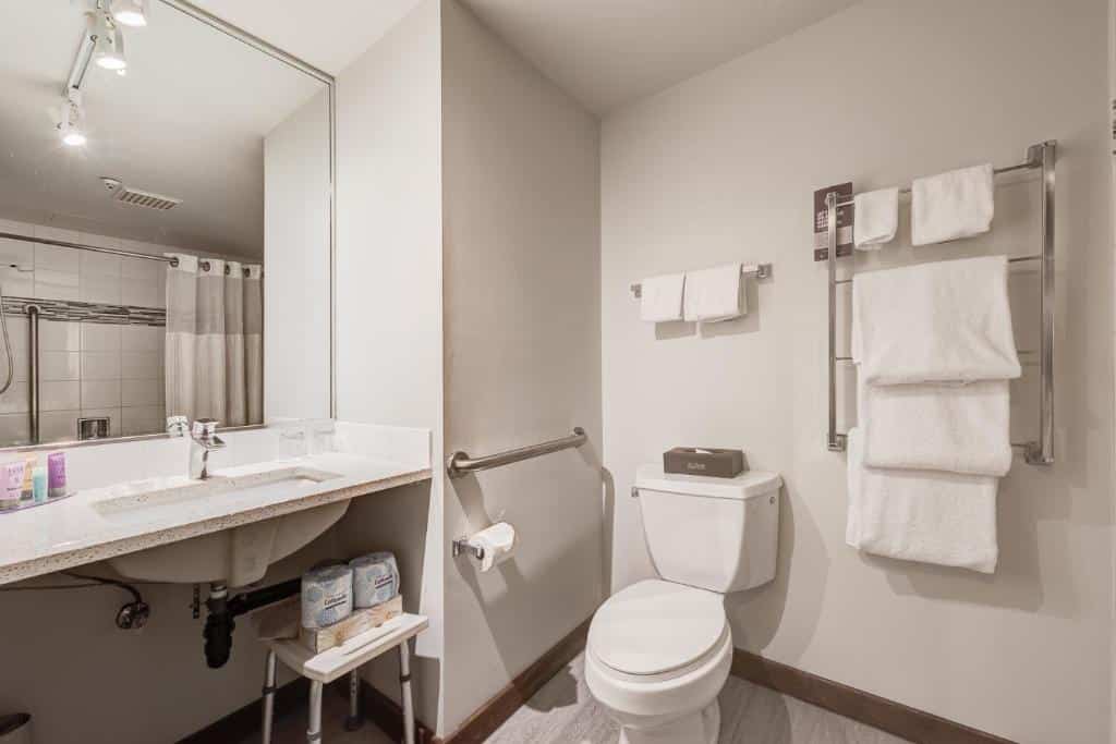 Banheiro adaptado do Blackcomb Springs Suites by CLIQUE com pia mais baixa e sem móvel embaixo, barras de apoio, um vaso sanitário e muitas toalhas ao dispor