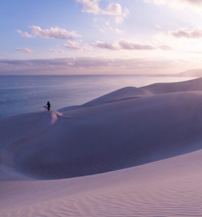 Dunas de areias brancas perto do mar no Iêmen. Há uma mulher com uma canga aberta ao longe, e o sol brilha no céu azul. - Foto: Andrew Svk via Unsplash