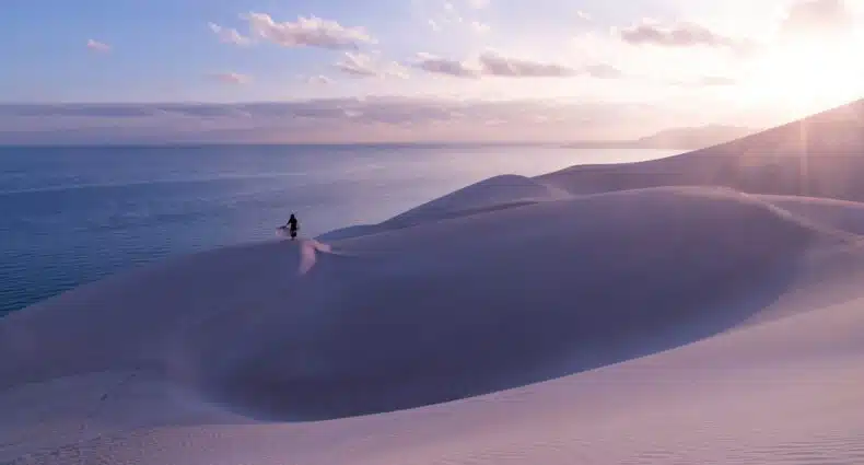 Dunas de areias brancas perto do mar no Iêmen. Há uma mulher com uma canga aberta ao longe, e o sol brilha no céu azul. - Foto: Andrew Svk via Unsplash