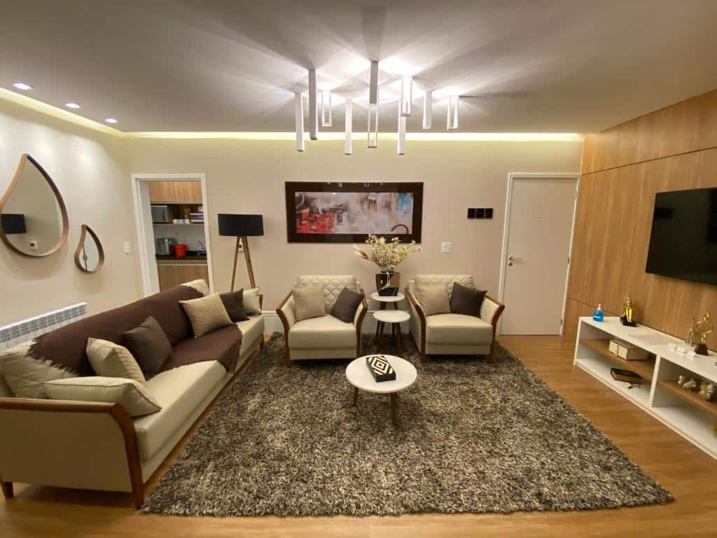 Sala de estar ampla do Apartamento em Campos do Jordão ao lado do Capivari com chão de madeira, tapete, sofás e poltronas, lustres, abajur de chão, televisão e espelhos