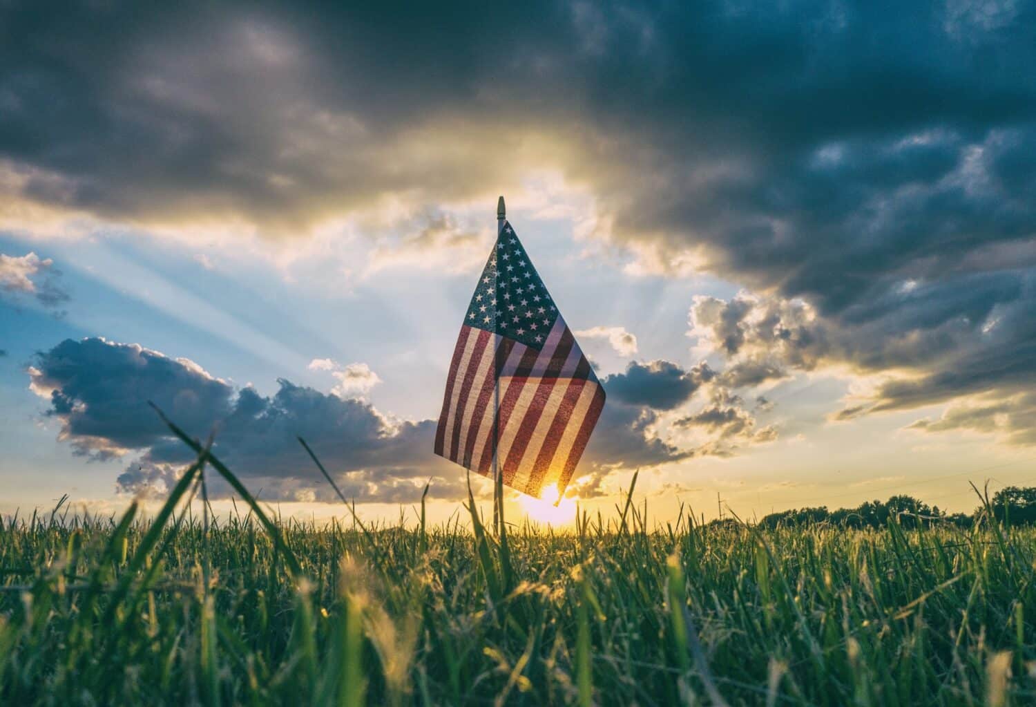 Bandeira dos EUA em haste fixada em um extenso gramado, em um dia de céu claro, com nuvens escuras e sol coberto por elas, para ilustrar a capa do post sobre como preencher o DS-160