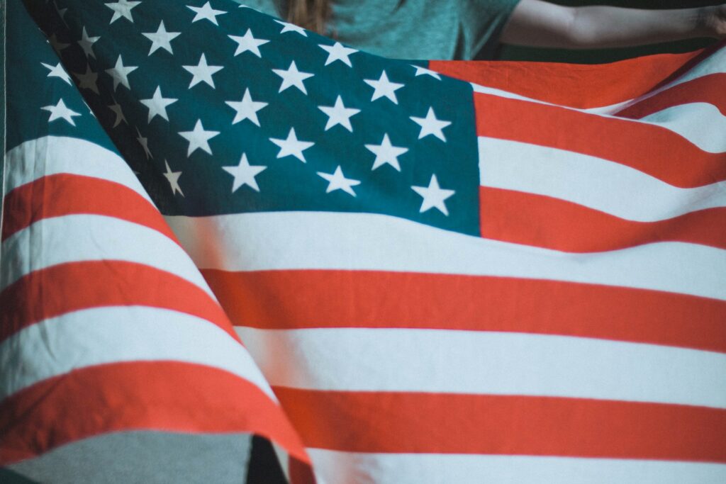 Bandeira dos Estados Unidos, grande, com listras vermelhas e brancas e estrelas brancas sobre um retângulo azul ao canto esquerdo, estendida por uma pessoa cujo braço é visto ao fundo