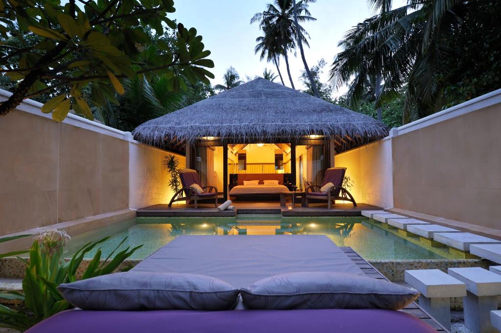 bangalô do Coco Bodu Hithi, com piscina e um colchão com almofadas do lado de fora e vista da varanda com cadeiras e da cama grande ao fundo