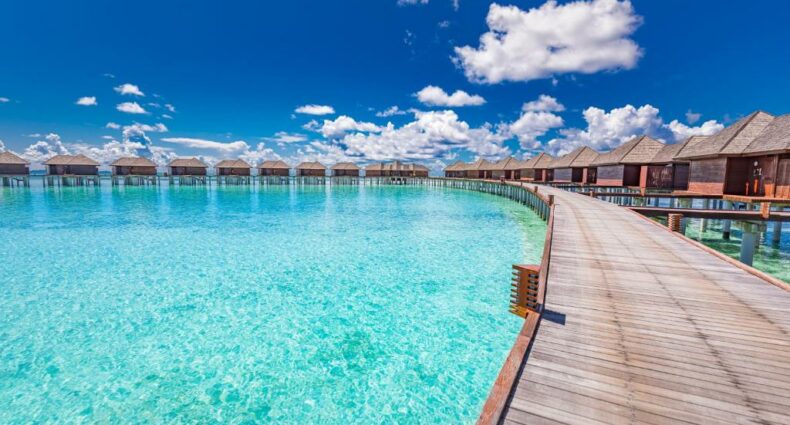 vista dos bangalôs com uma ponte de madeira comprida ligando cada um deles no Sun Siyam Olhuveli, um dos resorts nas Maldivas, com águas cristalinas abaixo e céu muito azul