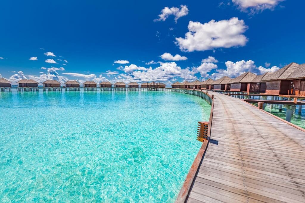 vista dos bangalôs com uma ponte de madeira comprida ligando cada um deles no Sun Siyam Olhuveli, um dos resorts nas Maldivas, com águas cristalinas abaixo e céu muito azul