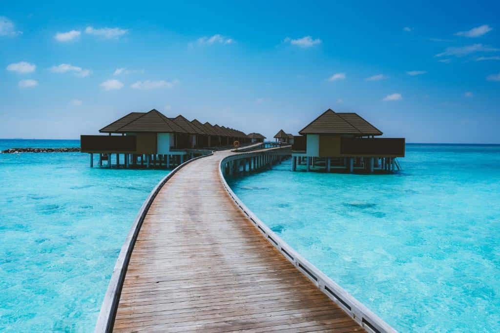 bangalôs iguais vistas de frente a partir da perspectiva de uma ponte de madeira com o mar abaixo e céu azul no VARU by Atmosphere, um dos resorts nas Maldivas