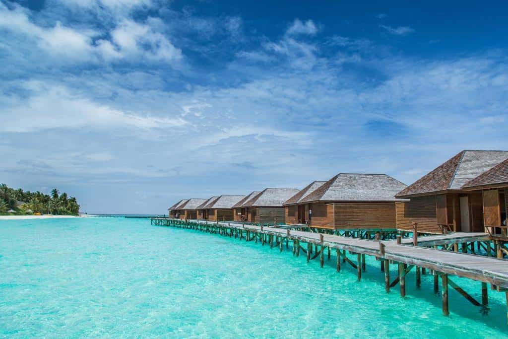 bangalôs iguais emm sequência, com uma ponte de madeira unindo cada um deles, no Veligandu Island Resort & Spa, um dos resorts nas Maldivas, o céu é azul com nuvens e o mar esverdeado e cristalino