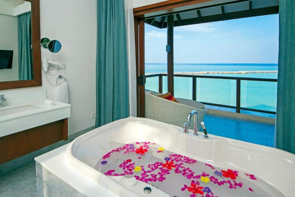 banheiro do Summer Island Maldives Resort, com banheira decorada com pétalas coloridas, pia grande de mármore e varanda com poltrona e vista para o mar