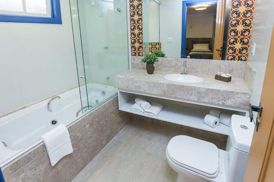 banheiro da Pousada Ouro Real com uma banheira à esquerda, um vaso sanitário à direita, e uma pia de pedra ampla com um grande espelho