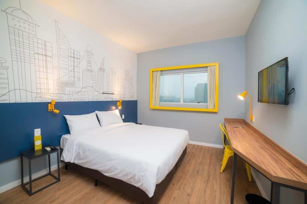 Quarto com parede desenhada no B&B Hotels São José dos Campos, além de mesinhas de cabeceira minimalistas, cama de casal com lençóis branquinhos, janela com cortina, mesa de trabalho e TV na parede