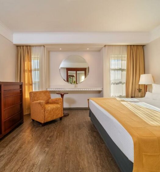 Quarto do Bourbon Atibaia Resort com cama de casal do lado direito com uma luminária do lado esquerdo da cama, em frente a cama com cômoda com TV e do lado esquerdo do quarto poltrona amarela. Representa resorts em Atibaia.