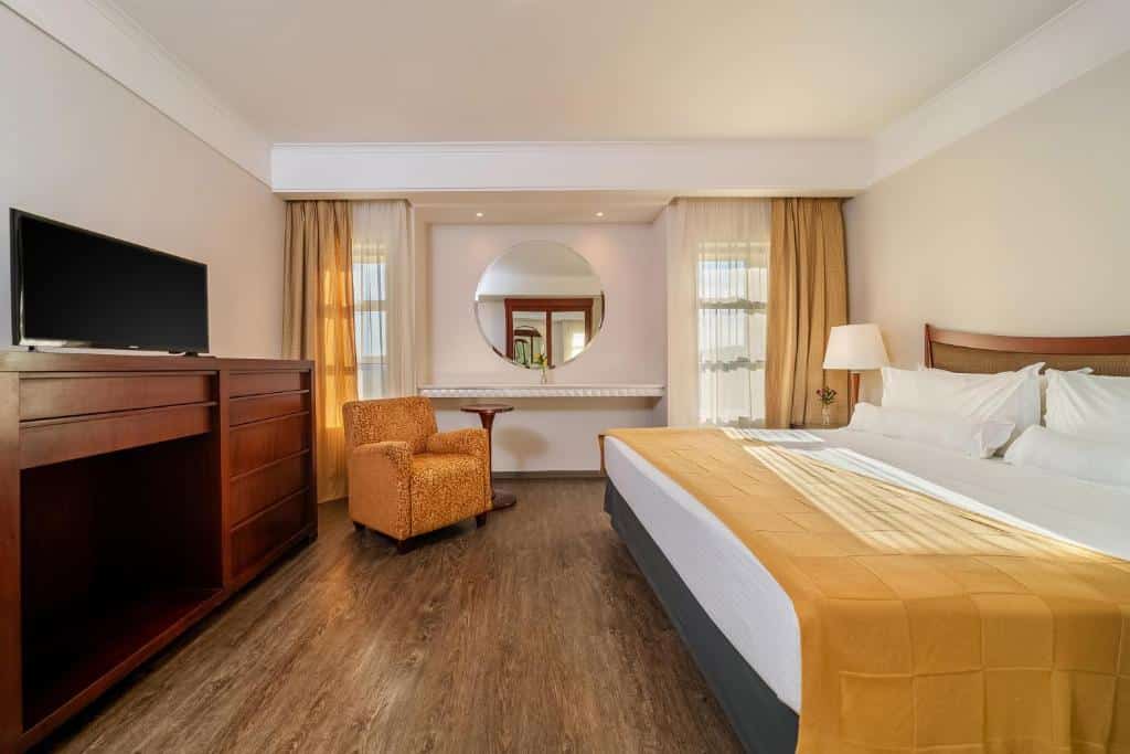 Quarto do Bourbon Atibaia Resort com cama de casal do lado direito com uma luminária do lado esquerdo da cama, em frente a cama com cômoda com TV e do lado esquerdo do quarto poltrona amarela. Representa resorts em Atibaia.