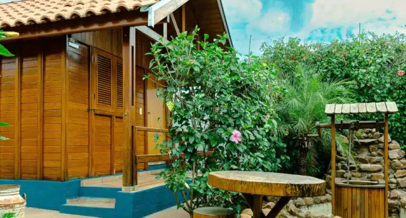 Frente da Pousada Recanto da Vó Ziza durante o dia, com estrutura rustica de madeira a hospedagem, em frente mesa de madeira e um poço. Representa chalés em Brotas.