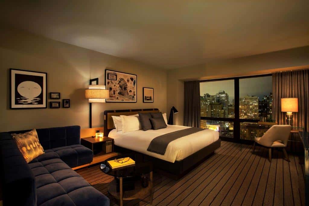 Vista do quarto do Thompson Chicago com sofá azul do lado direito, no centro do quarto uma cama de casal com duas cômodas ao lado com luminária e do lado esquerdo janelas amplas. Representa hotéis em Chicago.