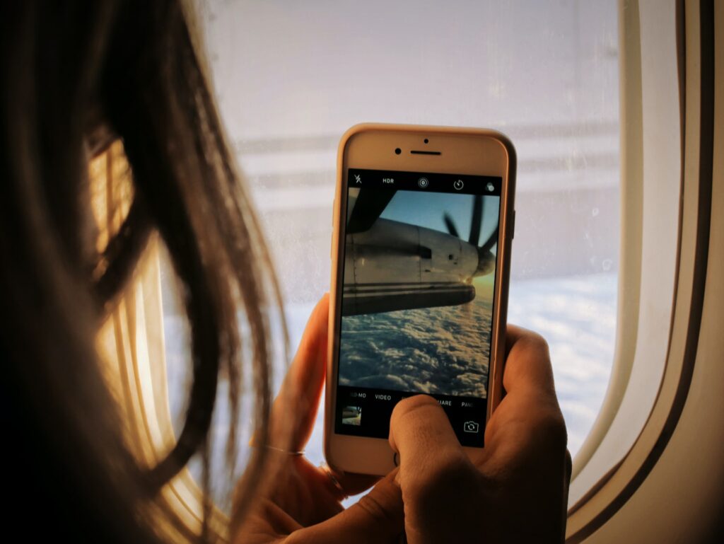 Mulher com cabelo longo segurando o celular com as duas mãos tirando foto pela janela do avião das nuvens do céu e da hélice do avião girando