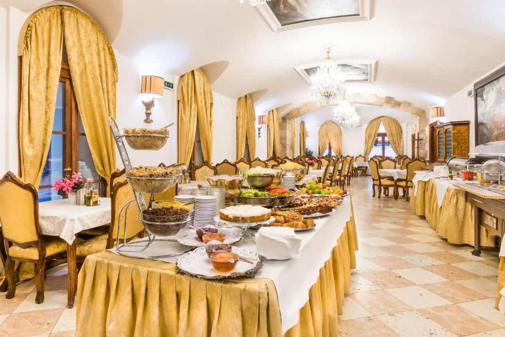 Sala de refeições do Charles Bridge Palace com mesas quadradas, cadeiras estofadas, janelas, lustres e tudo em tons de dourado