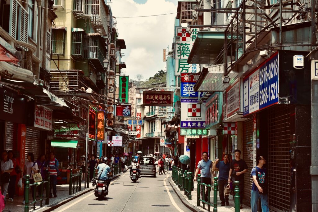 Rua em Macau, China, com pedestres nas calçadas ao lado, uma via ao meio por onde passam carros e motos e comércios locais ao entorno.