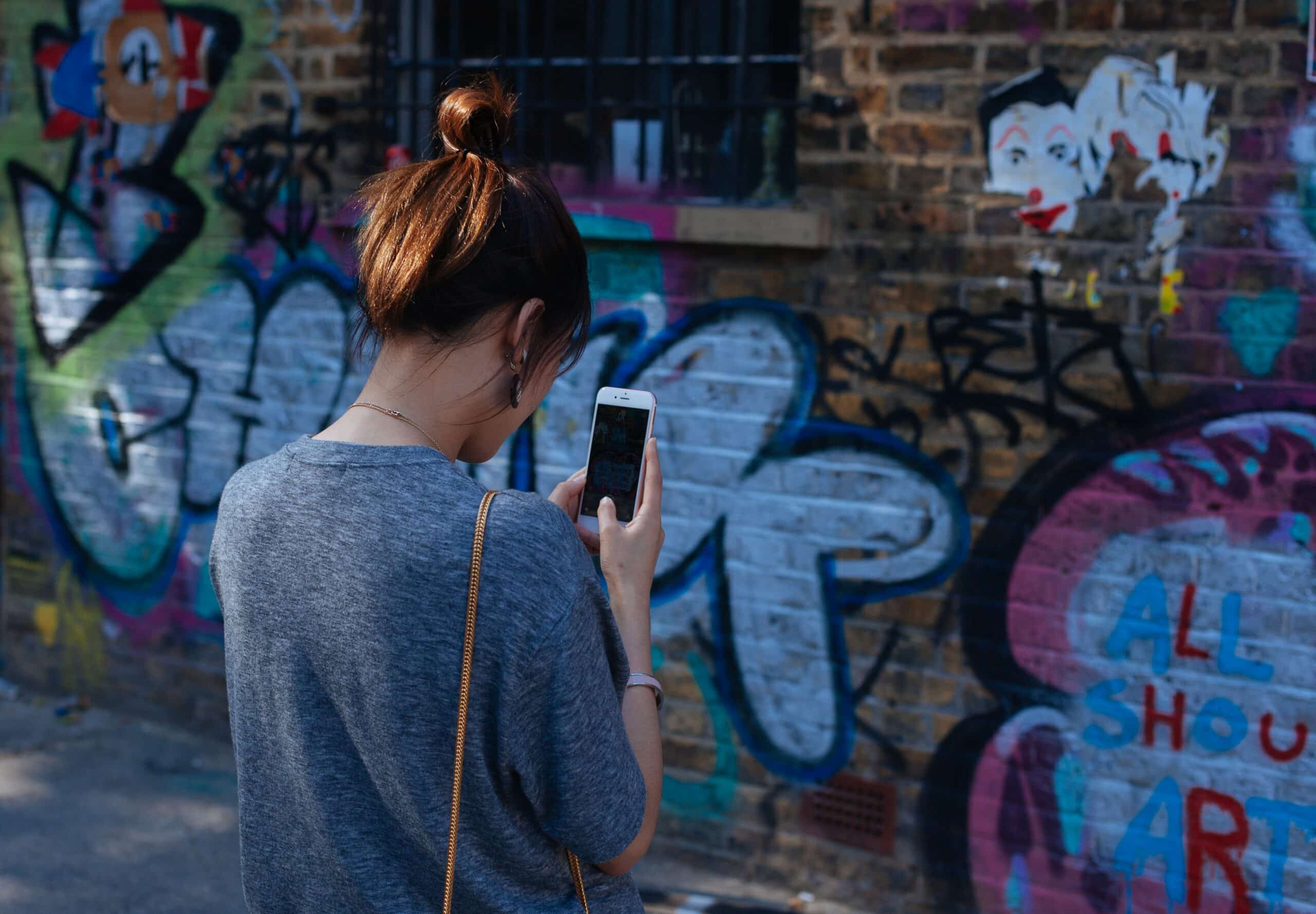 muro pichado de Brick Lane, na Inglaterra, com uma mulher de costas pra câmera, fotografando a parede. Ela é branca, cabelo liso e preso pra cima, usando um camisetão cinza e segurando um celular iphone branco para fotografar a arte. A imagem é usada para ilustrar como funciona o chip virtual para celulares compatíveis com o e-sim