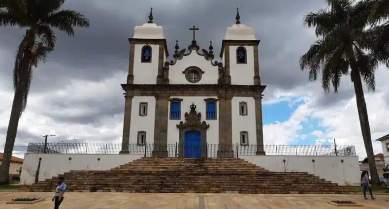 vista externa frontal da Igreja Matriz de Nossa Senhora da Conceição em Congonhas. De cada lado da igreja barroca há uma grande palmeira. Em frente há uma praça com vários degraus de pedra que formam a escada de entrada. O portão da igreja é azul, as paredes brancas e há duas torres, uma de cada lado da porta, com sinos