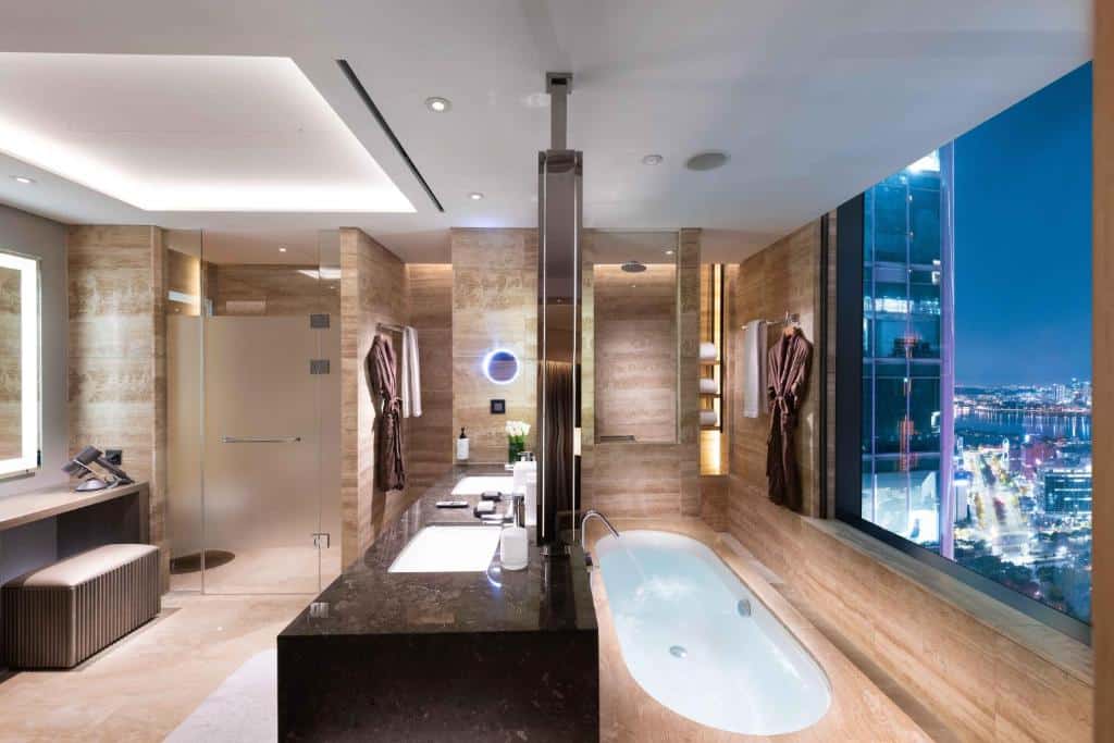 Banheiro do Conrad Seoul com uma janela ampla dando vista para a cidade, uma banheira de hidromassagem, pia com duas cubas e um box de vidro