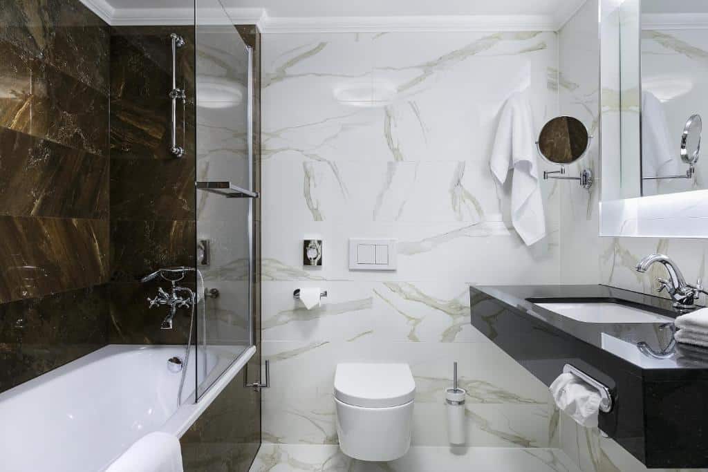 Banheiro do COSMOPOLITAN Hotel Prague com banheira de hidromassagem, um vaso sanitário, uma pia quadrada, um espelho amplo e algumas toalhas brancas pelo ambiente