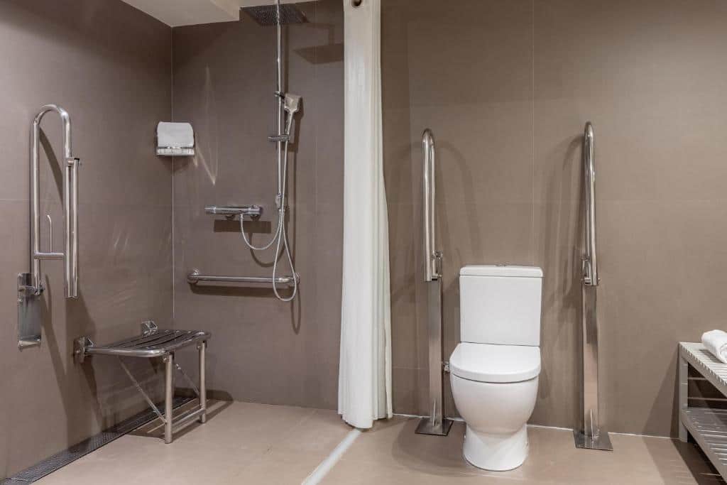 Banheiro adaptado do Duquesa Suites Barcelona, uma das recomendações de melhores hotéis em Barcelona com acessibilidade. Há barras de apoio e cadeira de banho perto do chuveiro, assim como barras de apoio ao lado da privada. Há uma cortina de banho branca ao lado do chuveiro.