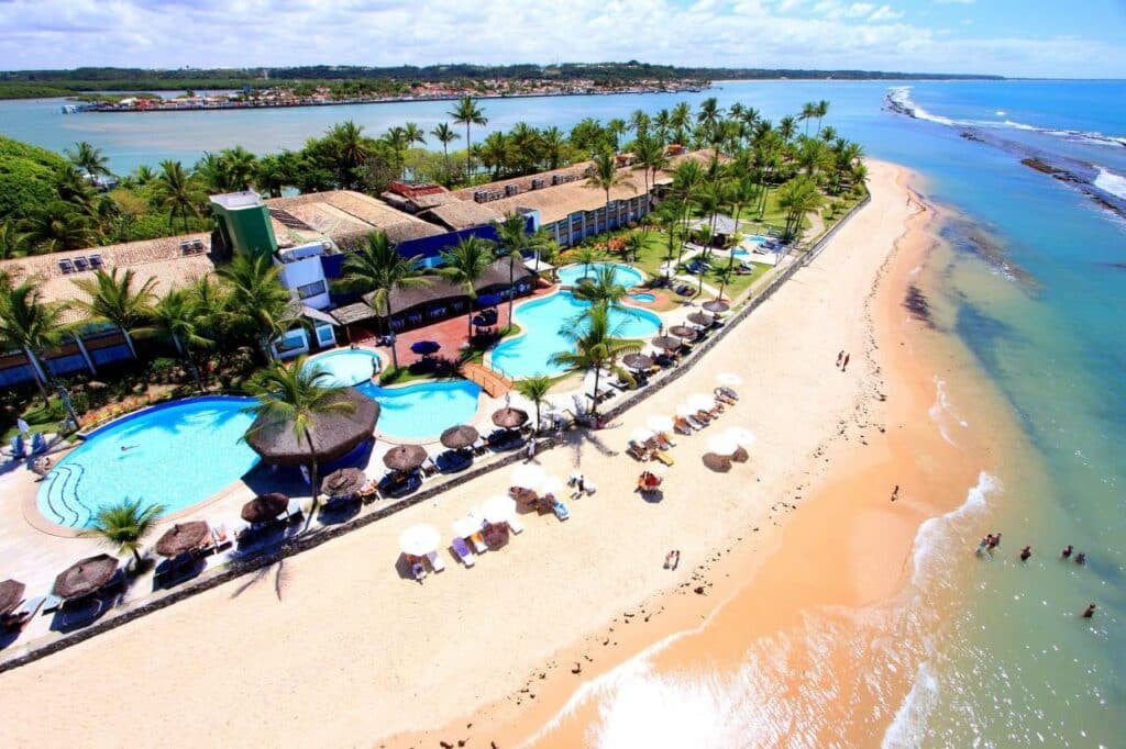Vista aérea do Arraial D'ajuda Eco Resort. A propriedade fica ao longo da praia, e a faixa de areia é bem em frente. Há várias piscina e árvores na frente do resort.