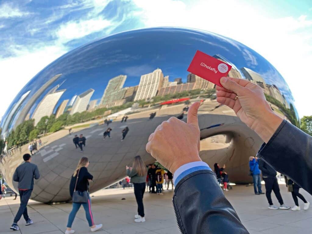 Uma mão segurando um cartão da Holafly e outra fazendo um sinal de jóia com a escultura "O Feijão", em Chicago, de fundo. A escultura é cromada prateada e reflete a imagem de prédios e da praça com pessoas andando
