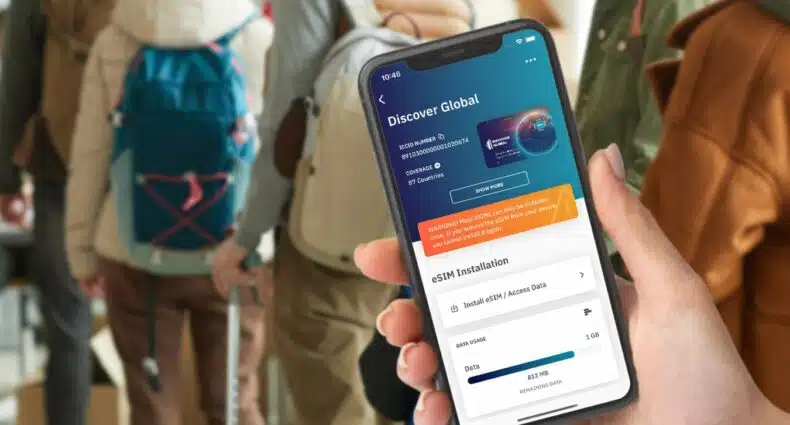 Mão segurando um celular com a tela mostrando a instalação de um plano da Airalo, e várias pessoas em uma fila com mochila nas costas ao fundo