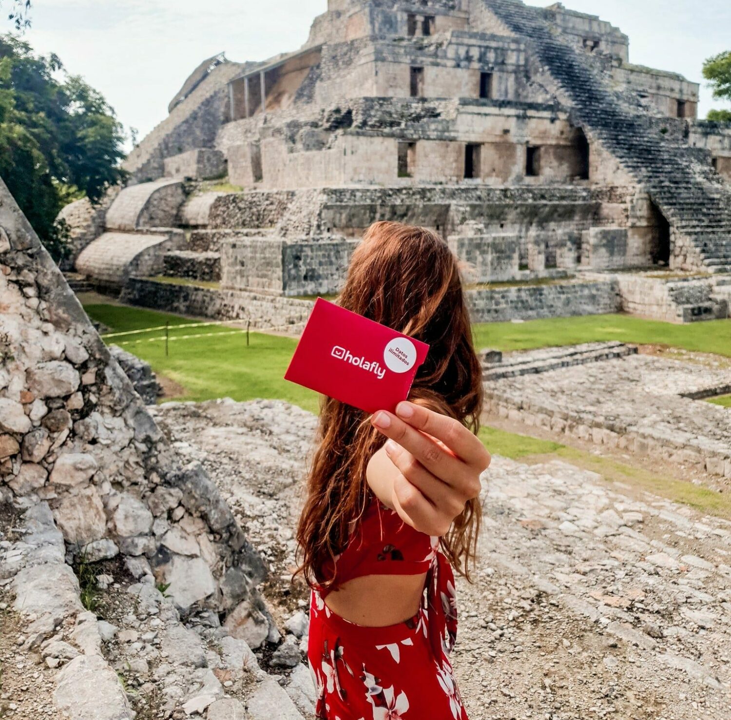 Mulher de cabelo longo, de costas, segurando um cartão do eSIM Holafly e olhando para uma pirâmide maia em Campeche, com gramado verde em volta e construções históricas