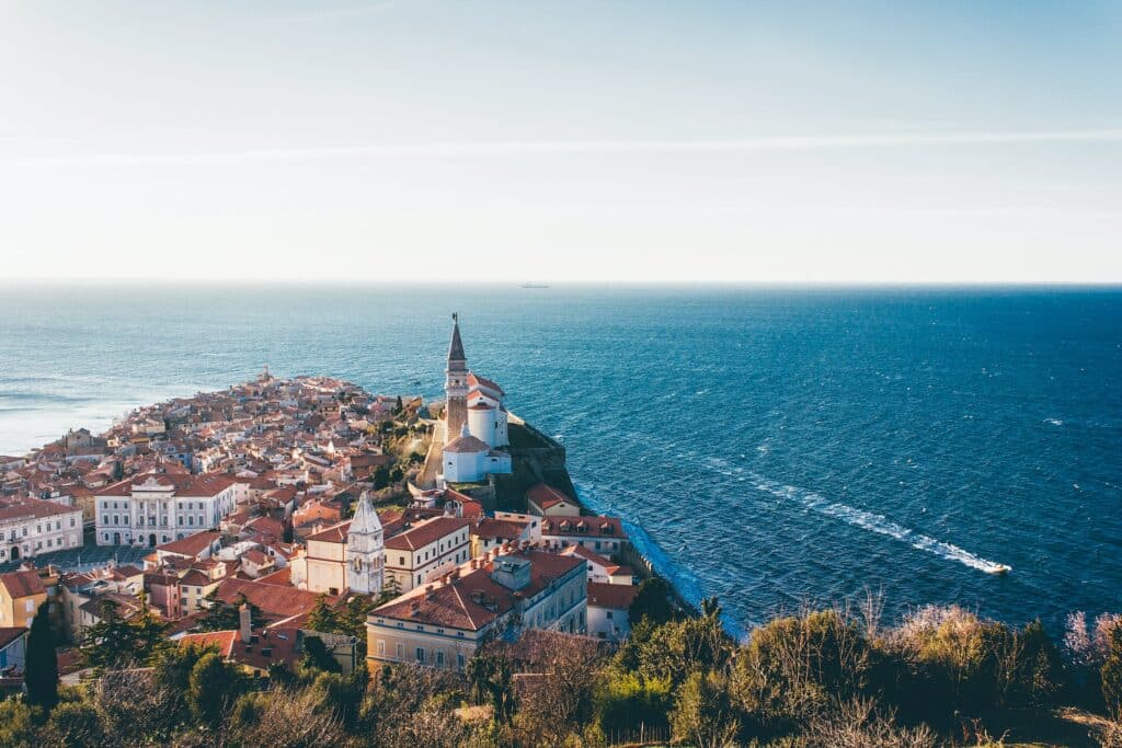Piran na Eslovênia, construções perto do mar azul cristalino com uma lancha passando em um dia de céu azul.