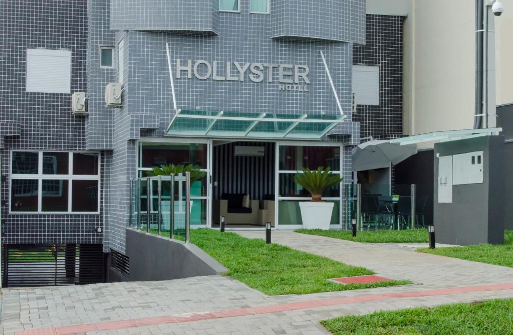 fachada do Hollyster Hotel, um dos hotéis perto do Aeroporto de Curitiba com cores cinza e o nome escrita na frente, há grama e uma garagem