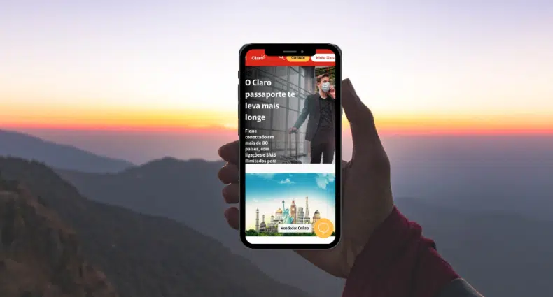 mão de uma pessoa segurando um celular com o print da página inicial do Passaporte Claro em detalhes vermelhos ao pôr do sol em meio a montanhas