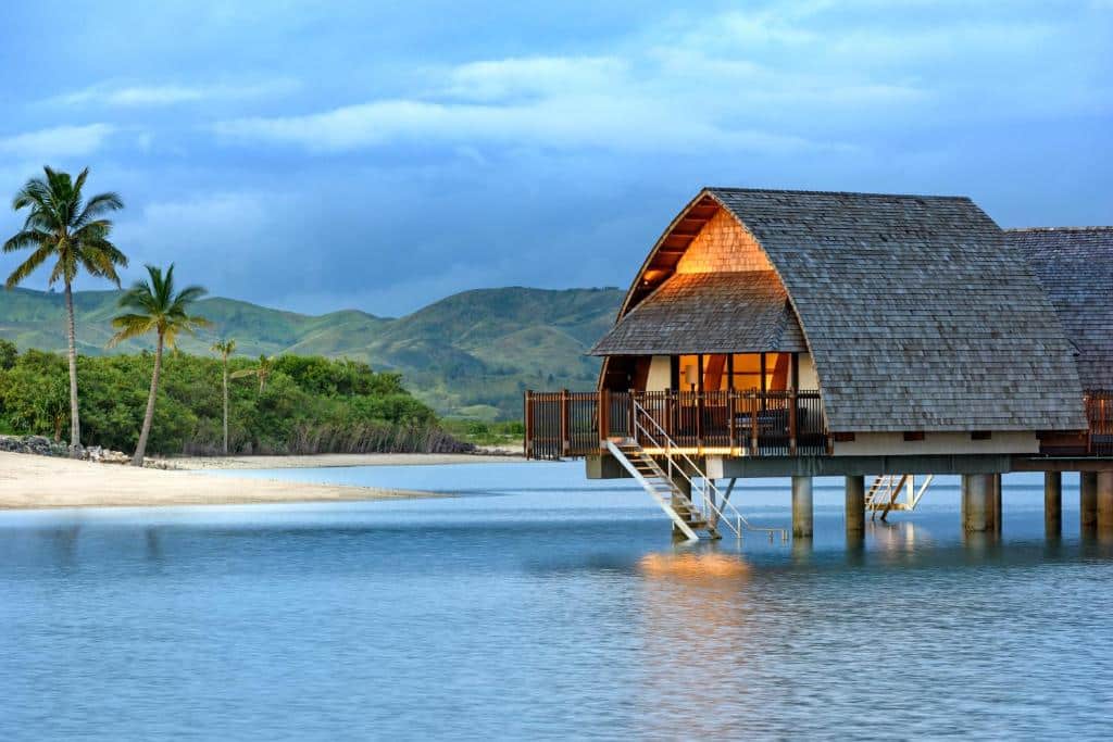 Frente do bangalô do Fiji Marriott Resort Momi Bay no final do dia, a acomodação fica suspensa dentro do mar com estrutura rústica. Representa hotéis em Fiji.