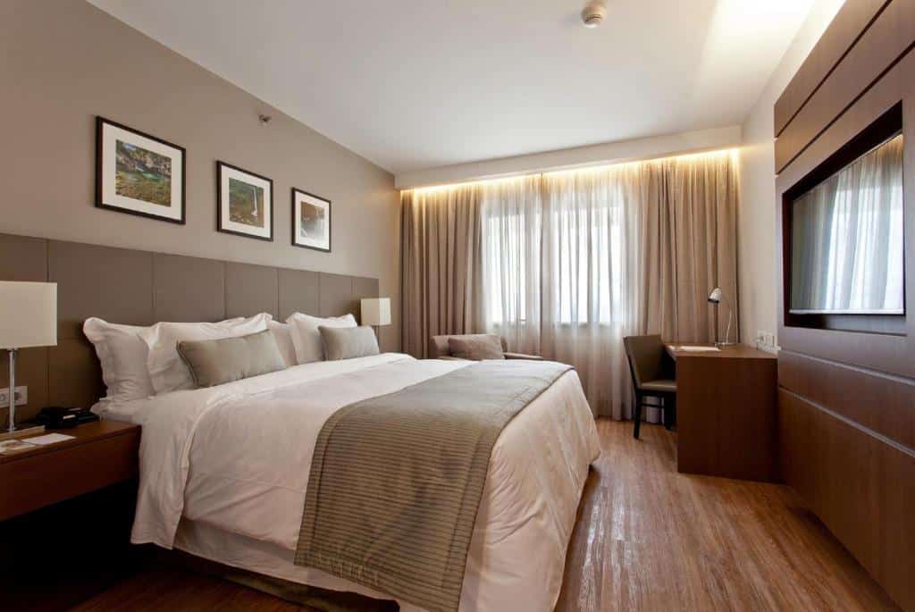 Quarto em um dos hotéis em São José dos Campos, o Golden Tulip, com cama de casal, TV grande, cortina dupla, mesa de trabalho, mesinhas de cabeceira com abajures, e três quatros decorativos sobre a cama.
